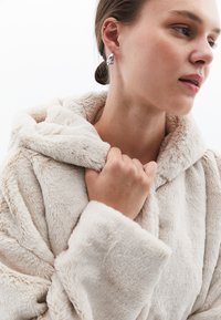 Kremowy kaptur z sztucznego futra o miękkiej texture, z kapturem, ściąganymi rękawami i dłonią trzymającą kołnierz.