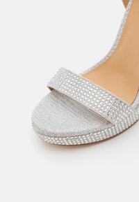 Sandale argentée à bout ouvert avec une large bride ornée de petits strass, un tissu texturé et une semelle plate et solide.