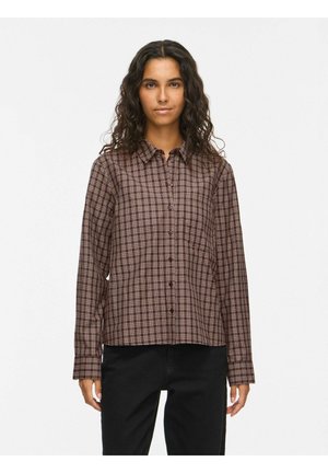 Femme aux longs cheveux bouclés portant une chemise à carreaux marron et blanche boutonnée et un pantalon noir, debout devant un fond uni.