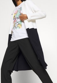 Lungo cappotto in maglia bianco e blu navy, con un motivo a texture. Sotto, una maglietta bianca con una grafica colorata. Pantaloni neri.