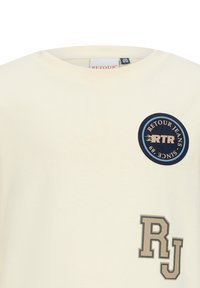 T-shirt w kolorze kremowym z okrągłym granatowym logo z napisem "RTR" oraz tekstem "RETLOUR JEANS SINCE 1989", a także grafiką "RJ" w jasnobrązowym odcieniu.