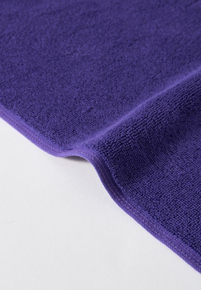 Μαλακό μωβ πανί μικροϊνών με υφή στην επιφάνεια και προσεγμένα ραμμένες άκρες, μερικώς διπλωμένο πάνω σε λευκό φόντο.