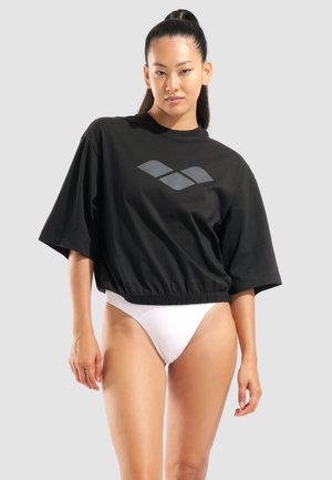 Kobieta z długimi ciemnymi włosami spiętymi w wysoki kucyk, ubrana w czarną, oversize, krótką koszulkę z logo oraz białe, wysokie bikini na szarym tle.