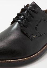 Scarpa nera in pelle con punta rotonda, superficie testurizzata, chiusura con lacci e dettagli marroni a contrasto sulla linguetta e sulla suola.