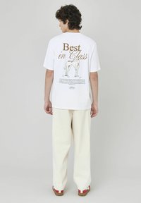 T-shirt en coton blanc avec un texte graphique marron "Meilleur de sa catégorie" et une illustration d'un chien au dos ; porté avec un pantalon blanc et des chaussures à accents rouges.