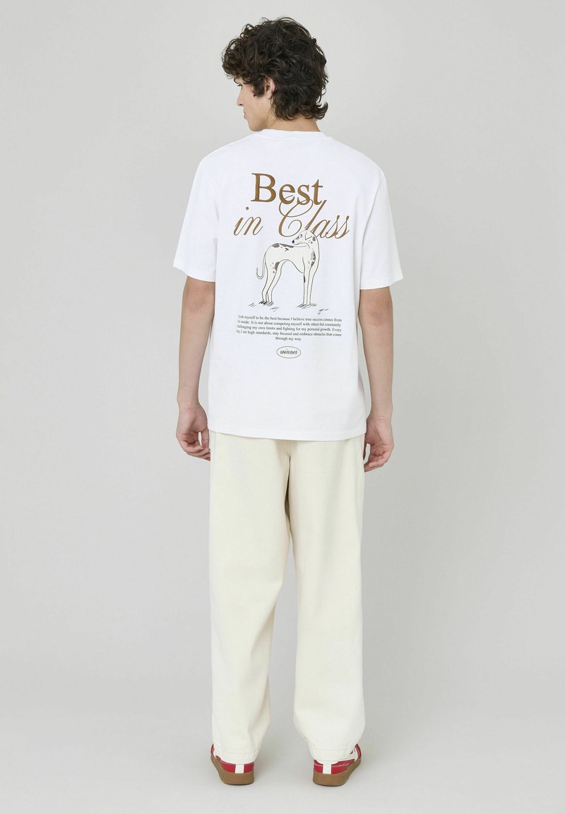T-shirt en coton blanc avec un texte graphique marron "Meilleur de sa catégorie" et une illustration d'un chien au dos ; porté avec un pantalon blanc et des chaussures à accents rouges.