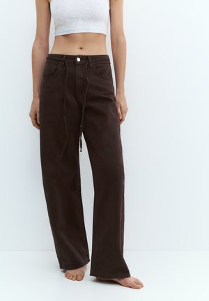 Kvinde iført løse mørkebrune bukser med høj talje og snøre, samt en hvid cropped ærmeløs top, stående barfodet op ad en hvid væg.