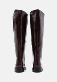 Augstas, burgundijas krāsas ādas zābaki ar strukturētu rakstu, ar sānu rāvējslēdzējiem un melnu plakanu zoli. Gluda apdare un pieguļošs dizains.