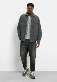 Gray wool blend jacket with two chest pockets, layered over a light green button-up shirt and gray T-shirt, paired with dark jeans and sneakers.