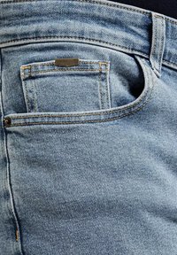 Jasnoniebieskie dżinsy z tylną kieszenią z małą metalową metką, widocznym szwem i teksturowaną powierzchnią materiału.