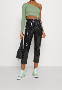 Frau trägt glänzende schwarze cropped Pants, ein grünes einärmliges Crop-Top, mintgrüne High-Top-Sneaker und hält eine kleine schwarze Tasche.
