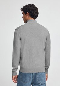 Grå stickad tröja med hög krage, långa ärmar och ribbade mudd och fåll. Baksidan har en slät yta och minimal detaljering.