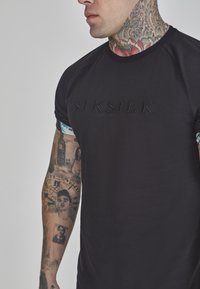 Melnā īsajām piedurknēm T-krekls ar reljefu "SIKSILK" logo priekšpusē, ar pieguļošu dizainu un teksturētu audumu. Redzamas tetovējumi uz rokas.