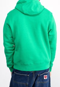 Sudadera verde con capucha y corte relajado, hecha de una tela suave. Cuenta con un bolsillo tipo canguro y puños acanalados, combinada con pantalones de mezclilla.