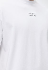 Λευκό φούτερ με στρογγυλή λαιμόκοψη και την επιγραφή "LIBERTINE LIBERTINE" εκτυπωμένη δύο φορές με μαύρο στο στήθος, φορεμένο από ένα άτομο.