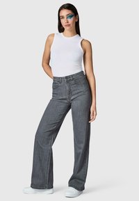 Femme debout, main dans le dos, portant un top blanc sans manches, un jean gris taille haute à jambes larges, des chaussures blanches et un maquillage bleu à l'œil gauche.