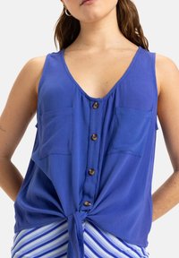 Blouse sans manches bleue avec un décolleté en V, des boutons à l'avant, deux poches poitrine et un détail de taille nouée. Tissu lisse avec une texture douce.