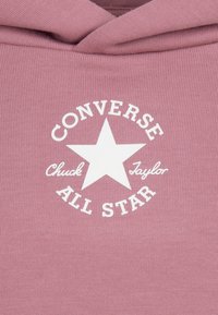 Puha textúrájú rózsaszín kapucnis pulóver fehér kör alakú logóval, amelyen csillag látható. A logó szövege: "Converse Chuck Taylor All Star."