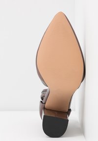 Chaussure en cuir marron à bout pointu avec une semelle lisse beige et un petit talon noir reposant sur une surface blanche.