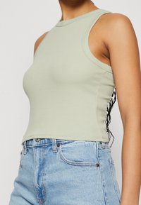 Světle zelený žebrovaný top bez rukávů s vysokým výstřihem. Má boční výřezy a přiléhavý střih. Kombinován s modrými džínovými šortkami.