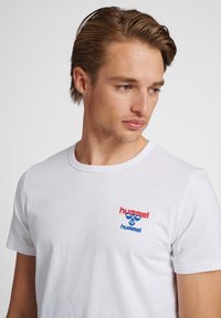 Młody mężczyzna o jasnobrązowych włosach, ubrany w białą koszulkę z czerwono-niebieskim logo Hummel, patrzy w dół i w prawo na jednolitym tle.