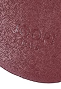 Rund, bordeaux läderyta präglad med "JOOP! JEANS" i ett slätt typsnitt, med en kantig textur.