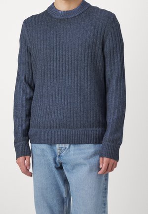 Mand iført mørkeblå ribstrikket sweater med rund hals og lyseblå cowboybukser, stående mod en ensfarvet baggrund.