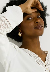 Blusa blanca con borde de encaje floral y escote con forma de concha. La modelo tiene el pelo corto y rizado y lleva pendientes de oro. Textura de piel suave.