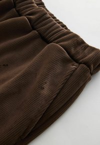 Bruine corduroy-stof met verticale ribbels, voorzien van een elastische tailleband en een zachte textuur, gedeeltelijk gedrapeerd om de details en de vorm te tonen.
