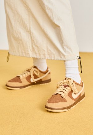 Personne portant des baskets Nike beiges et marron, des chaussettes blanches et un pantalon ample de couleur claire, debout sur un sol jaune.