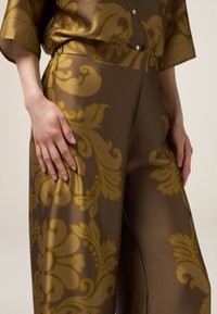 Braune Satin-Hosen haben ein großes goldfarbenes Blumenmuster, einen lockeren Schnitt und eine glatte Textur. Eine Hand liegt auf dem Stoff in der Nähe des Bundes.