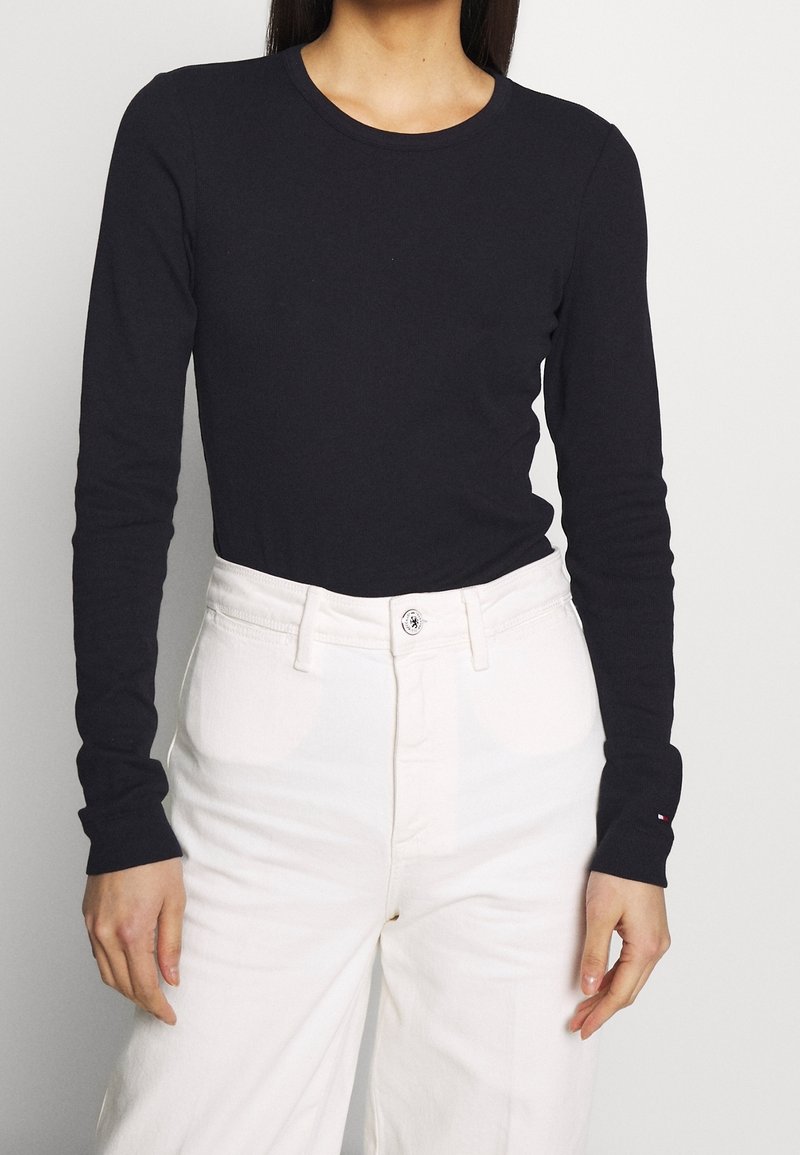 Femme portant un haut noir ajusté à manches longues, rentré dans un pantalon blanc taille haute à jambes larges, sur un fond uni.