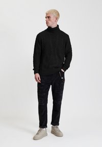 Zwarte gebreide coltrui met kabelpatroon, gecombineerd met zwarte versleten broek en lichtgrijze sneakers op een neutrale achtergrond.