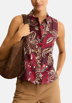 Donna con blusa floreale bordeaux senza maniche, pantaloni beige e borsa intrecciata marrone, una mano in tasca, su sfondo semplice.