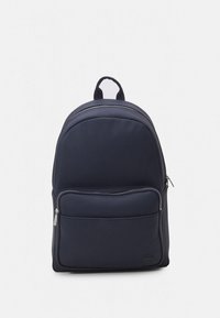 Mörkblå ryggsäck med en mjuk, texturerad yta; har en dragkedjeförsedd framficka och justerbara remmar. Lacoste-logotyp längst ner.