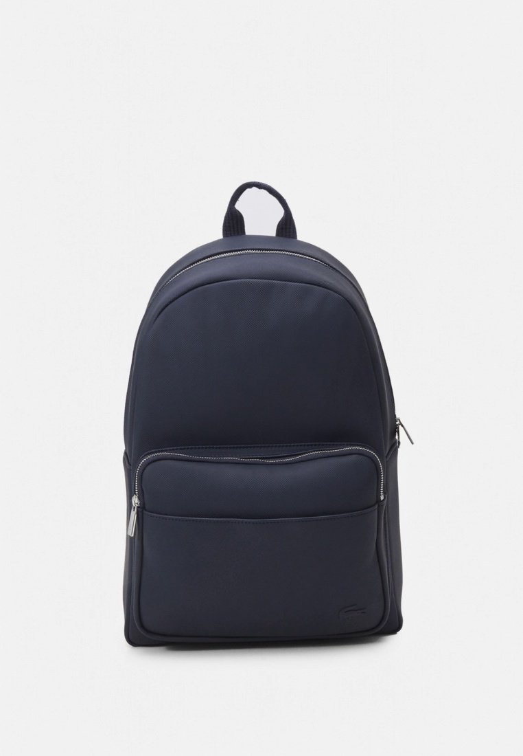 Sac à dos bleu marine avec une finition lisse et texturée ; comporte une poche avant zippée et des bretelles réglables. Logo Lacoste en bas.