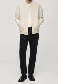 Mann trägt eine cremefarbene, geknöpfte Collegejacke, ein weißes Hemd, schwarze gerade Jeans und schwarze Lederschuhe und steht mit den Händen in den Taschen.