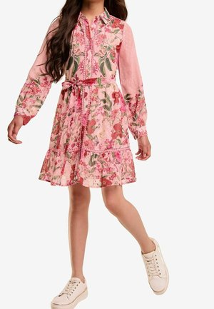 Rosa Blumenkleid mit langen Ärmeln, Knopfleiste vorne und gerafftem Rock. Abgerundet mit passendem Gürtel und getragen zu weißen Sneakern.