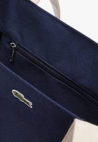 Sac fourre-tout en toile bleu marine avec une poche zippée sur le dessus, rehaussé d'un petit logo brodé, et poignées beiges pour le contraste et la durabilité.