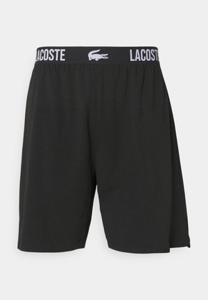 Schwarze Shorts mit elastischem Bund, die mit weißem "LACOSTE"-Text und Logo versehen sind. Aus glattem Stoff gefertigt, mit lockerer Passform und ohne Taschen.