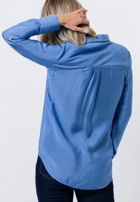 Camisa azul claro de manga larga, hecha de una tela suave. Presenta un cuello, detalles de botones en las mangas y un dobladillo redondeado.