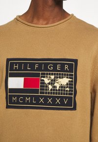 Beige Strickpullover mit einem marineblauen rechteckigen Patch mit dem Text "HILFIGER", einem roten Kasten und einer Weltkarte in goldener Stickerei.