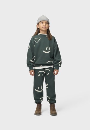 Barn iført mørkegrønt matchende sweatshirt og bukser med hvide smiley-ansigter, grå strikhue og brune støvler, stående mod en ensfarvet baggrund.