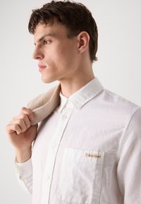 Camicia bianca con bottoni realizzata in tessuto testurizzato, dotata di taschino sul petto con il logo Calvin Klein. Un capo beige è appoggiato sulla spalla.