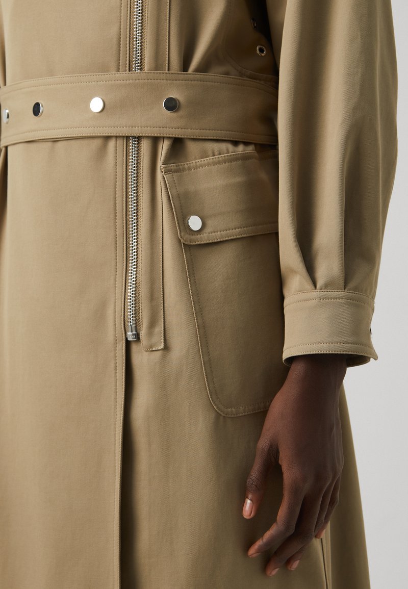 Beige, langärmlige Oberbekleidung mit einem Reißverschluss, Druckknopfverschlüssen und einer Seitentasche. Glatter, strukturiertes Material mit einer Taillengürtel-Detail.