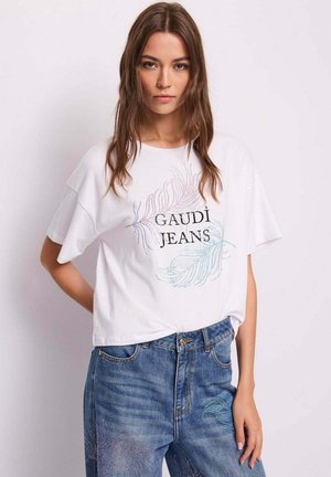 Donna che indossa una maglietta bianca Gaudi Jeans con motivi di piume e jeans blu a vita alta con decorazioni di piume abbinate.
