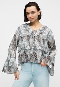 Jeune femme portant un chemisier ample à motifs avec des volants et des manches évasées, associé à un jean taille haute bleu clair, mains dans les poches.