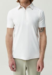 Hvit polo skjorte med en kort glidelås, myk tekstur, svak glans og et subtilt mønster nær kragen. Kombineres med lyse bukser.