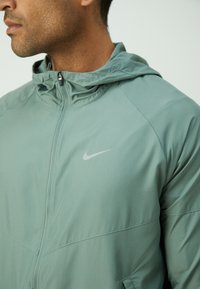 Veste Nike vert clair avec fermeture éclair à l'avant, dotée d'une capuche et d'un logo discret. Fabriquée en matériau lisse et léger avec une finition mate.