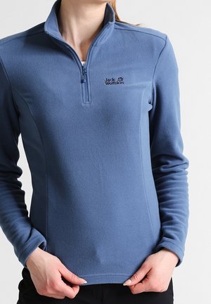 Kvinde iført en blå Jack Wolfskin fleece-pullover med kvart lynlås, der holder i kanten af pullovren med begge hænder.
